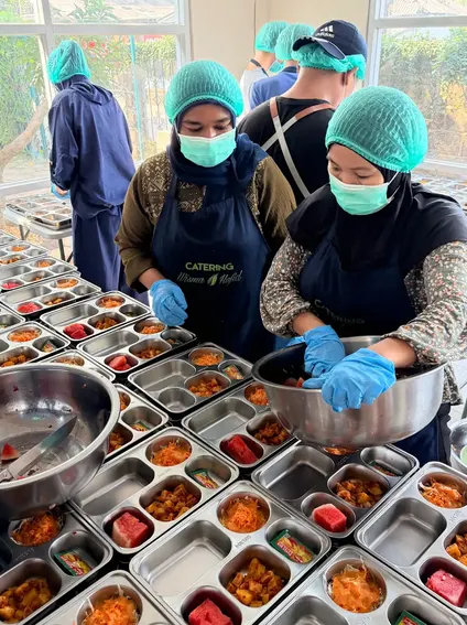 Beberapa staf menyiapkan dan mengisi tray makanan secara berurutan di dapur.