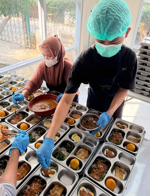 Staf bersarung tangan mengisi tray makanan di dapur.