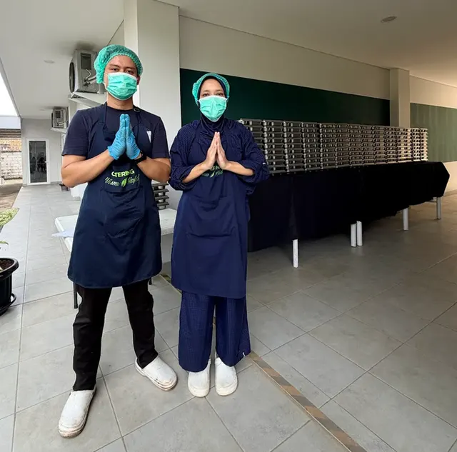 Dua staf Wisma Hafid Catering mengenakan celemek, penutup kepala, dan masker di area penyiapan.